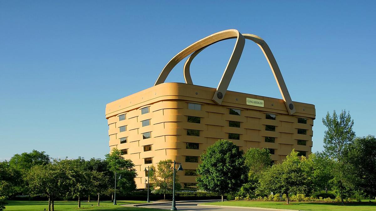 Image of Longaberger building in Newark Ohio on a clear cloudless day just before sunset with trees and the driveway in the foreground and the building in the background. A large, multi-story tan building designed to resemble a woven wicker basket with two handles leaning over its roof.